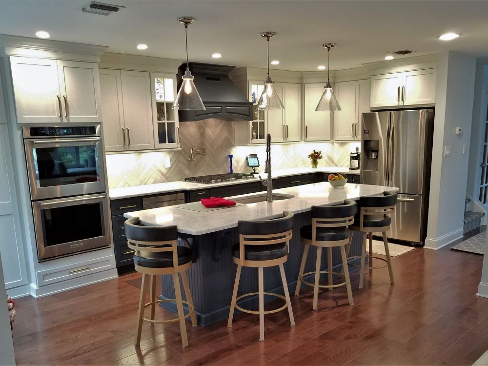 In this overview of the No. Haledon, NJ kitchen, the large island with seating anchors the space and is the focus of the room; family and friends can enjoy a bite while the host prepares food or cleans up. The two-toned painted Homecrest cabinetry keeps the kitchen interesting, while the wood flooring warms up the room.